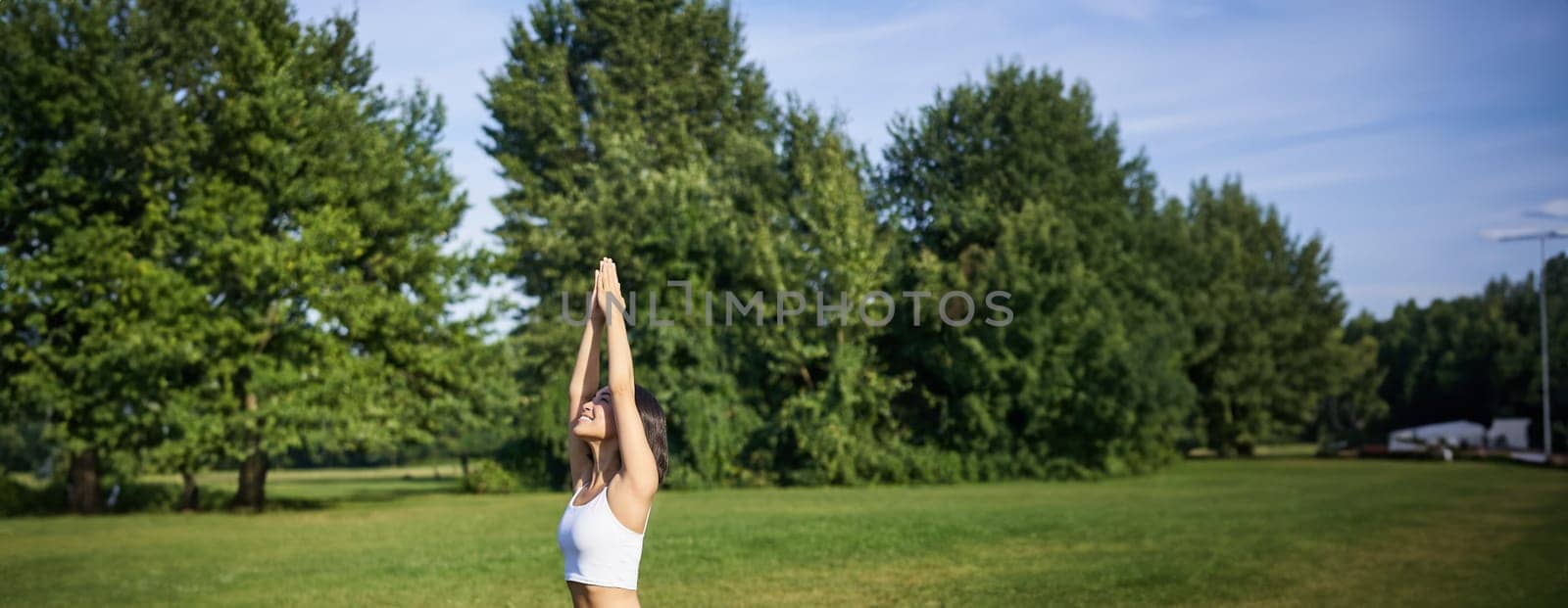 Vertical shot of smiling korean woman doing tree yoga asana, stretching on rubber mat in park, exercising.