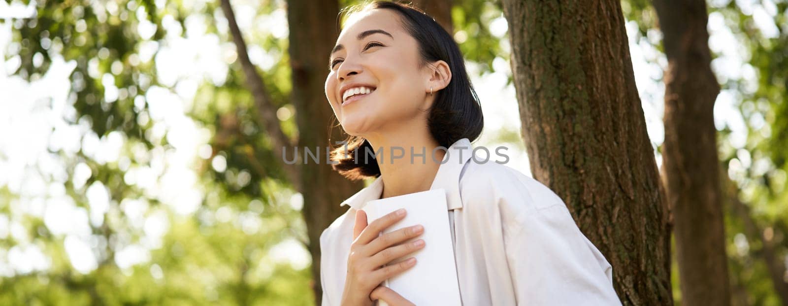 Romantic smiling girl reading book in park or foret, sitting under tree shade on sunny day, relaxing on fresh air surrounded by nature.