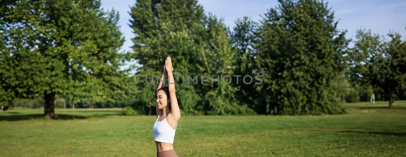 Vertical shot of smiling korean woman doing tree yoga asana, stretching on rubber mat in park, exercising.