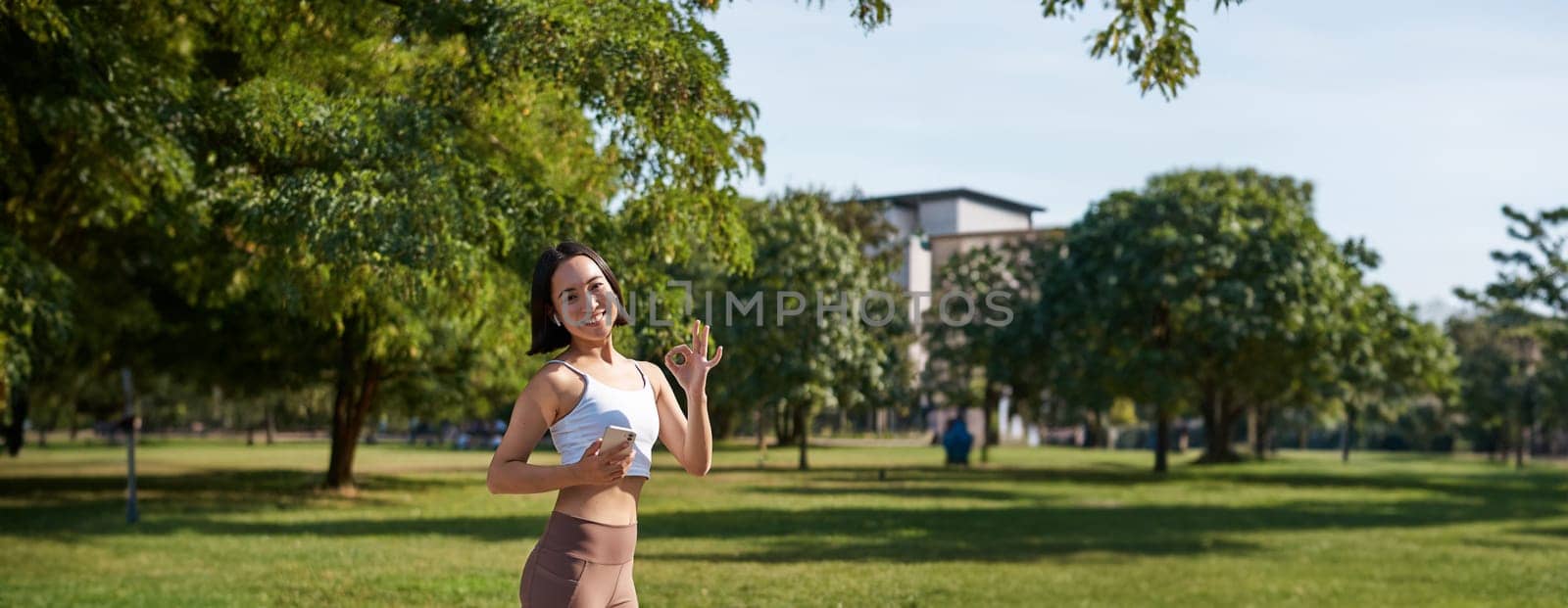 Sport and wellbeing. Young sportswoman in park, listen music and smiling, workout outdoors, jogging on streets of city.