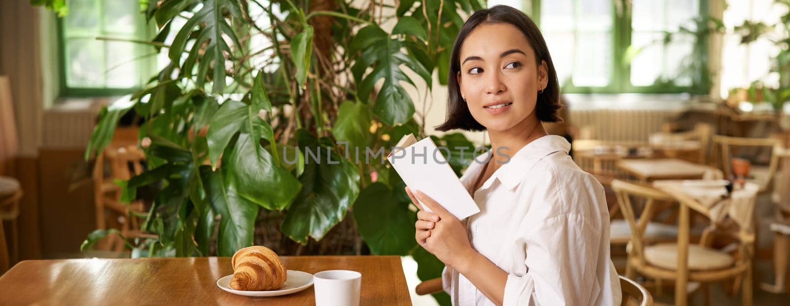 Beautiful young asian woman with a book in hands, sitting in cafe, drinking coffee and eating croissant, smiling, looking mysterious.