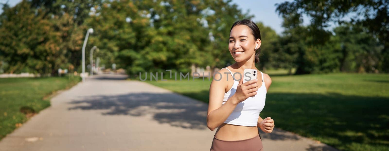 Happy smiling asian woman jogging in park. Healthy young female runner doing workout outdoors, running on streets.