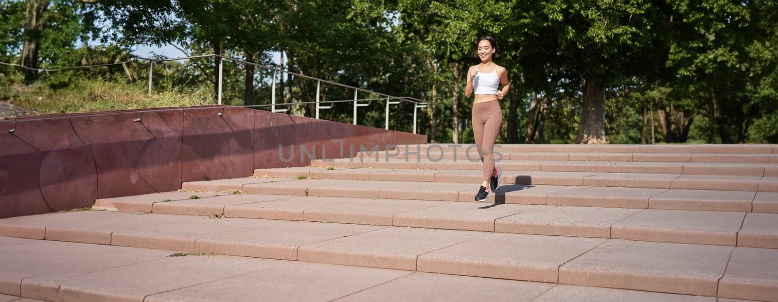 Portrait of young sportswoman running on street. Fitness girl jogging in park, workout outdoors, listening music in wireless earphones.