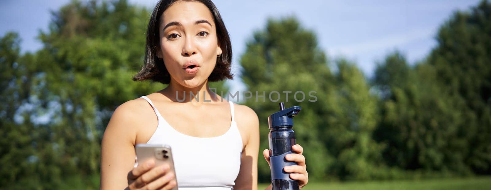 Excited young asian woman workout and drinks water, follows sports app on smartphone, looks amazed and happy, does excercises in park.
