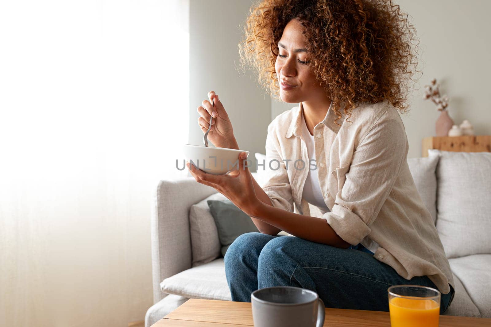 Young relaxed pensive African American woman eating healthy breakfast sitting on the couch. Copy space. by Hoverstock