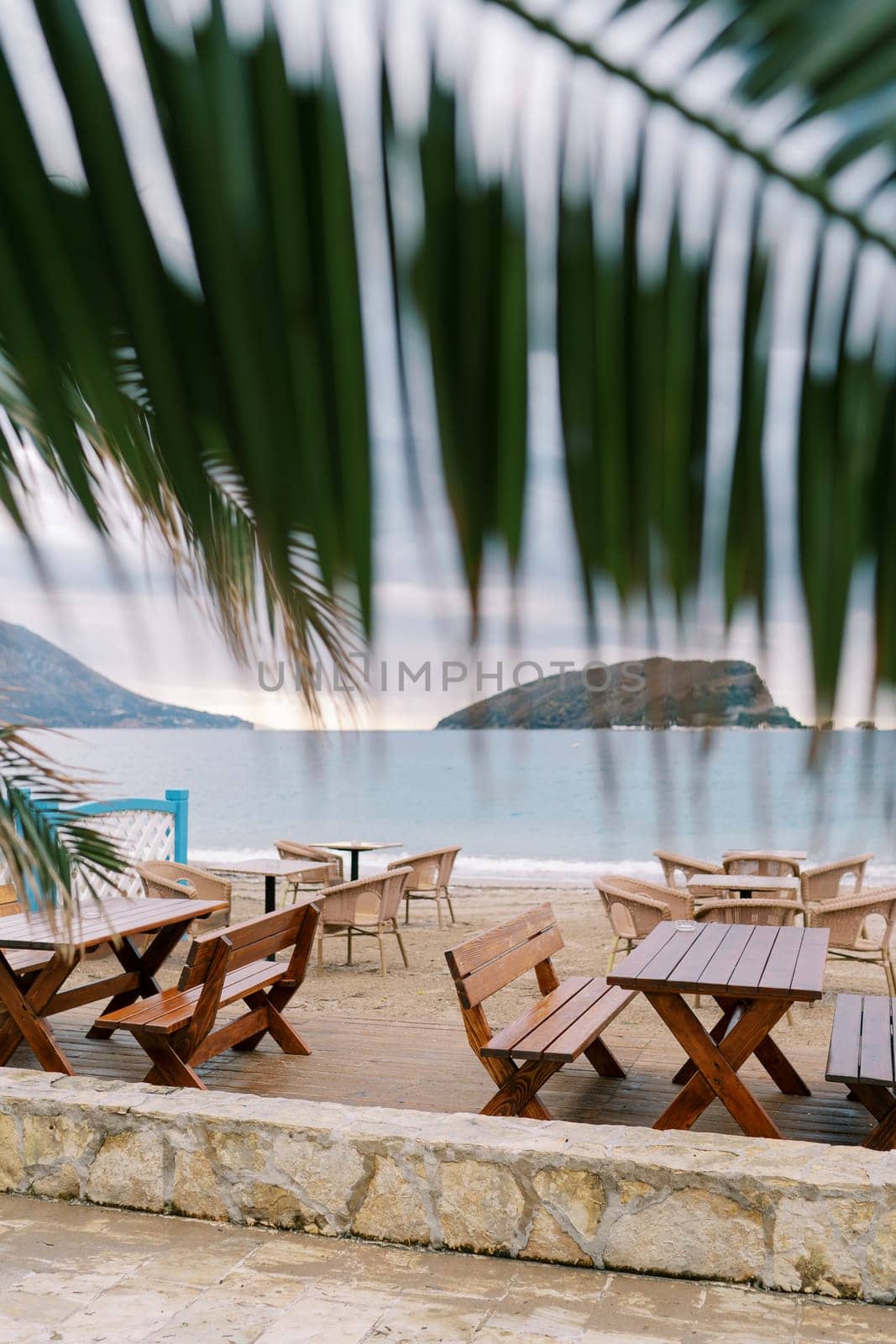 View from behind a green palm leaf on wooden tables and chairs standing on the beach by the sea by Nadtochiy