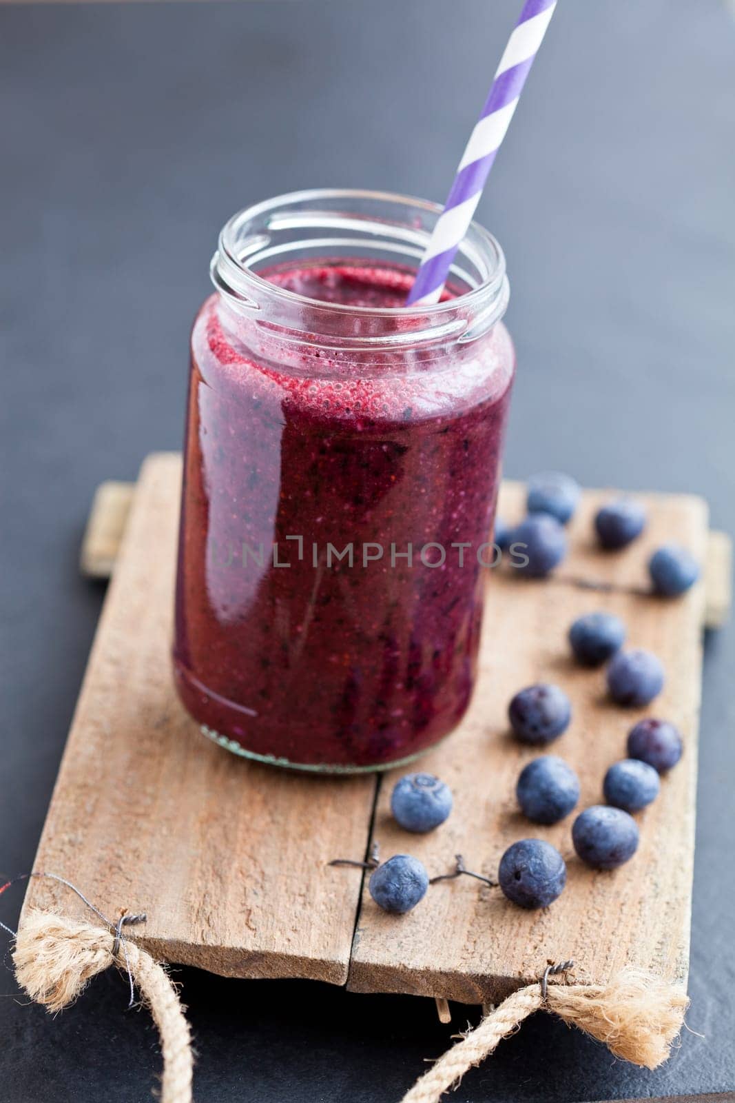 Closeup of blueberry smoothie in glass with striped straw on wooden serving tray with blueberries
