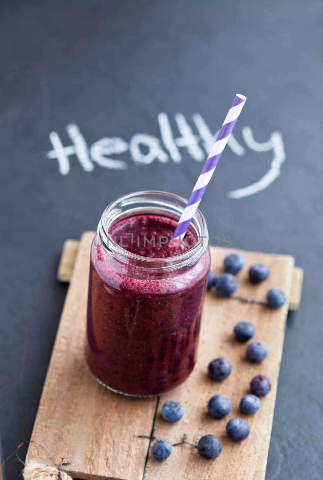 Closeup of blueberry smoothie in glass with striped straw on wooden serving tray with word healthy written on dark background