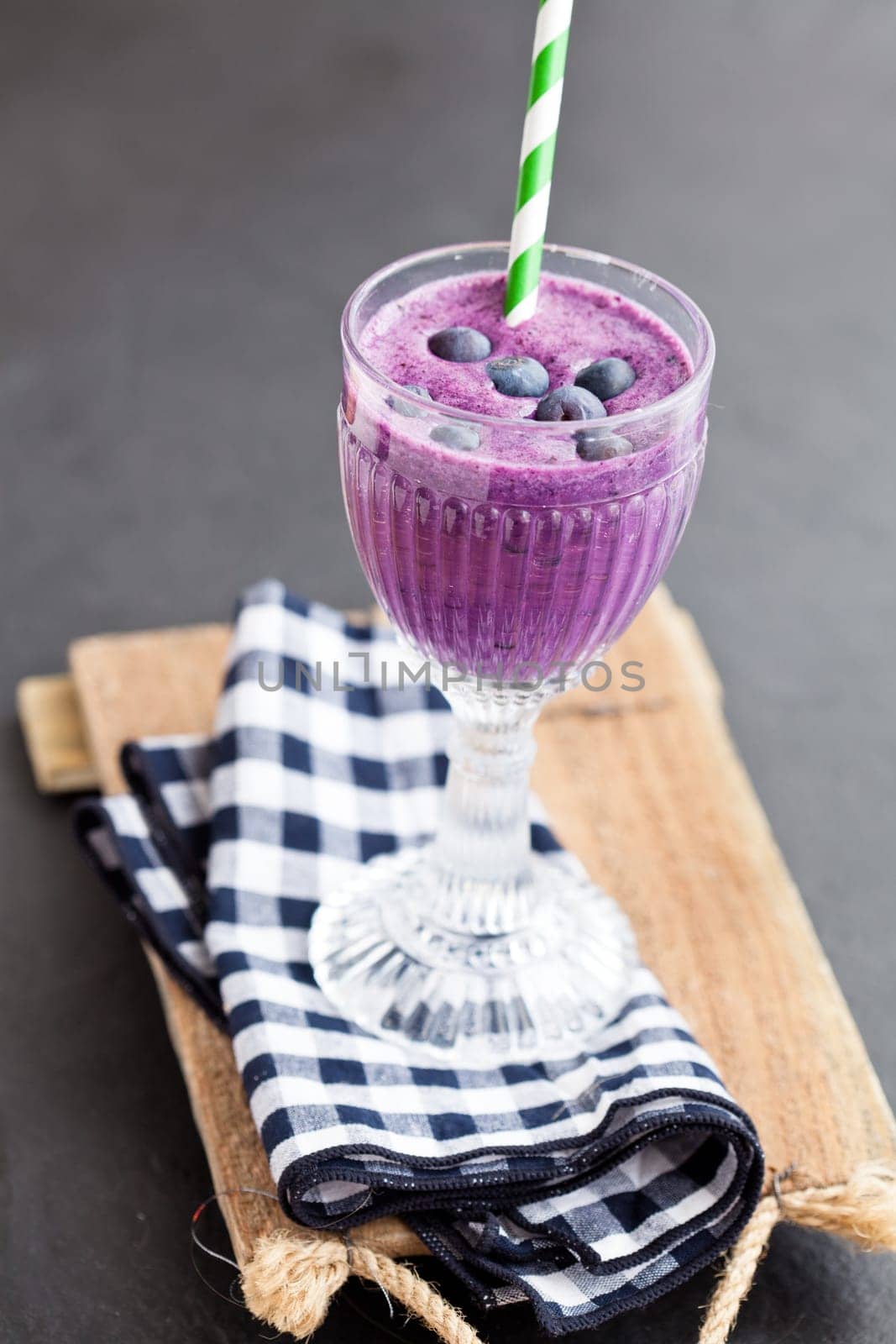 Closeup of blueberry milk smoothie in glass with striped straw on checkered napkin on wooden serving tray