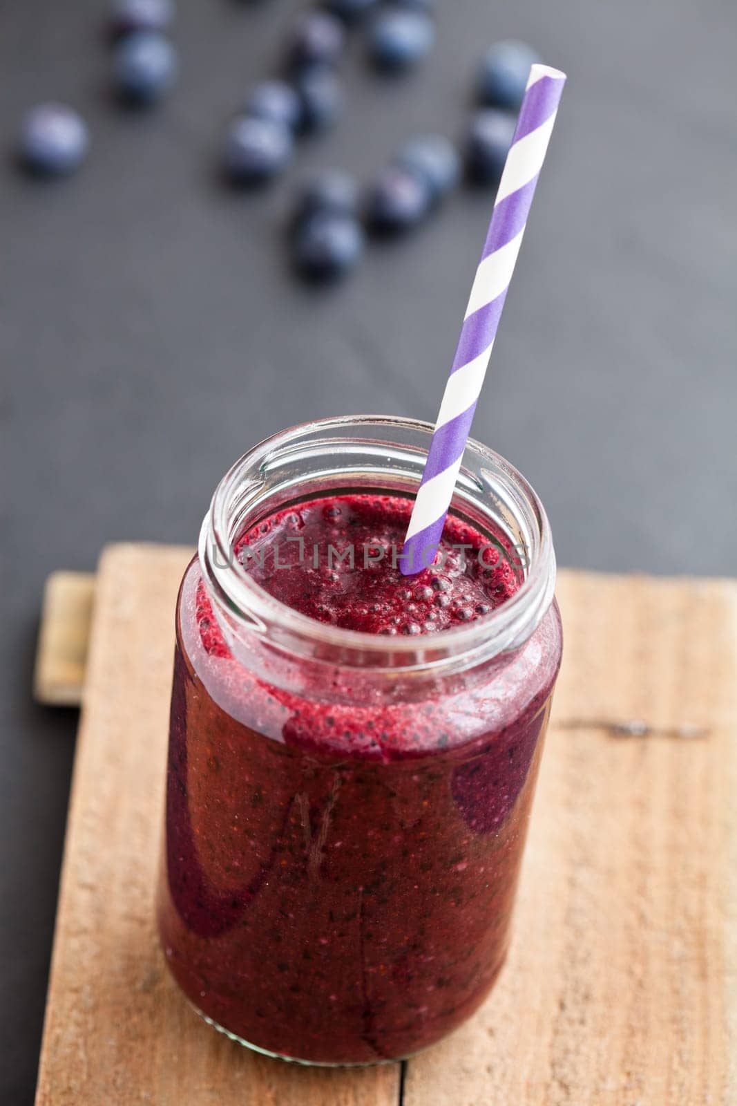 Closeup of blueberry smoothie in glass with striped straw on wooden serving tray with blueberries in background