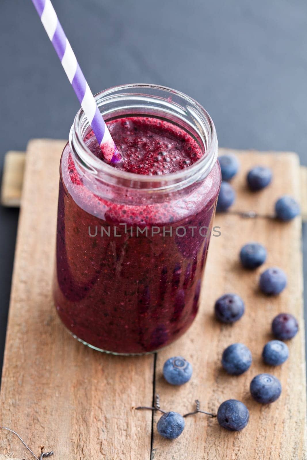 Closeup of blueberry smoothie in glass with striped straw on wooden serving tray with blueberries