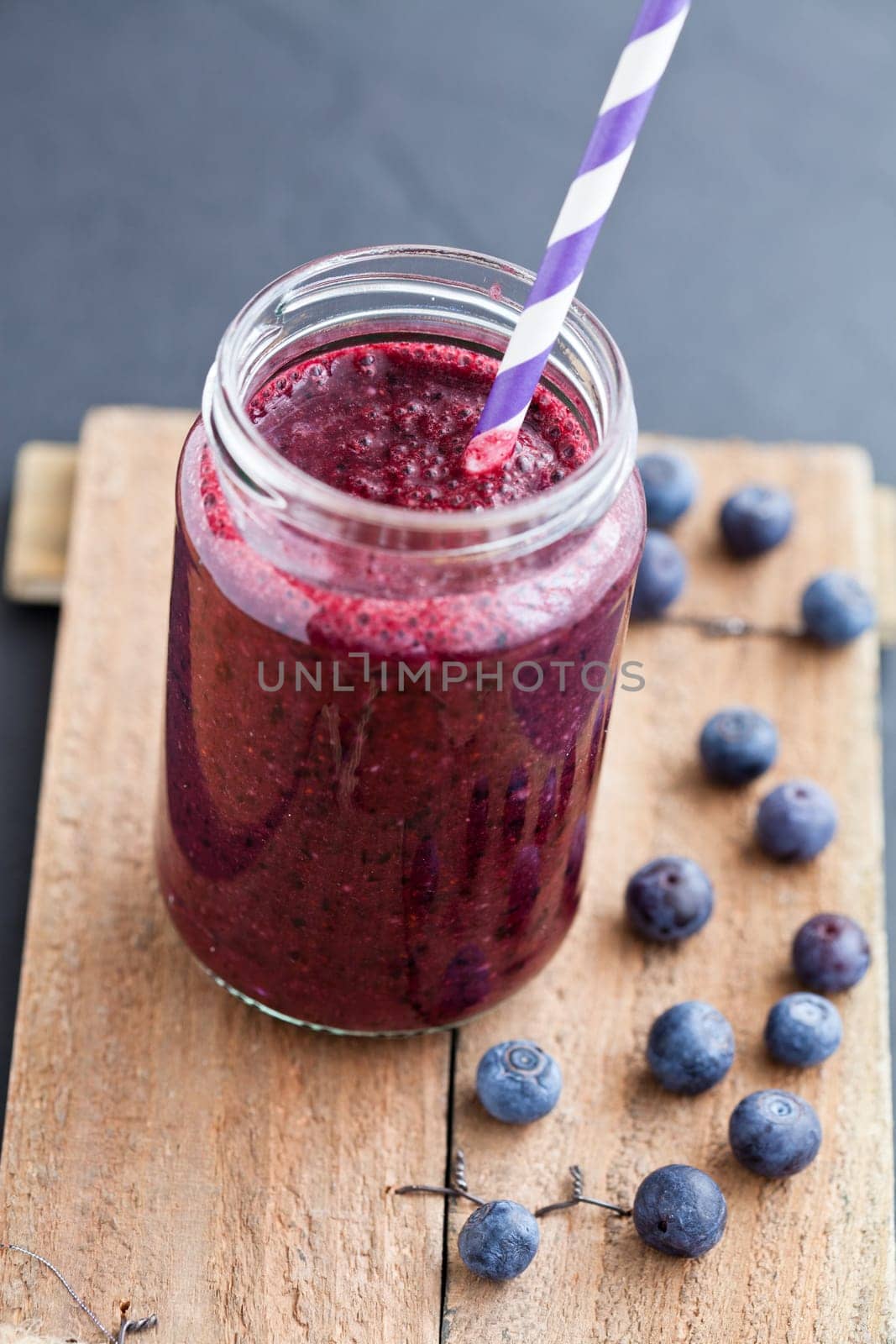 Closeup of blueberry smoothie in glass with striped straw on wooden serving tray with blueberries