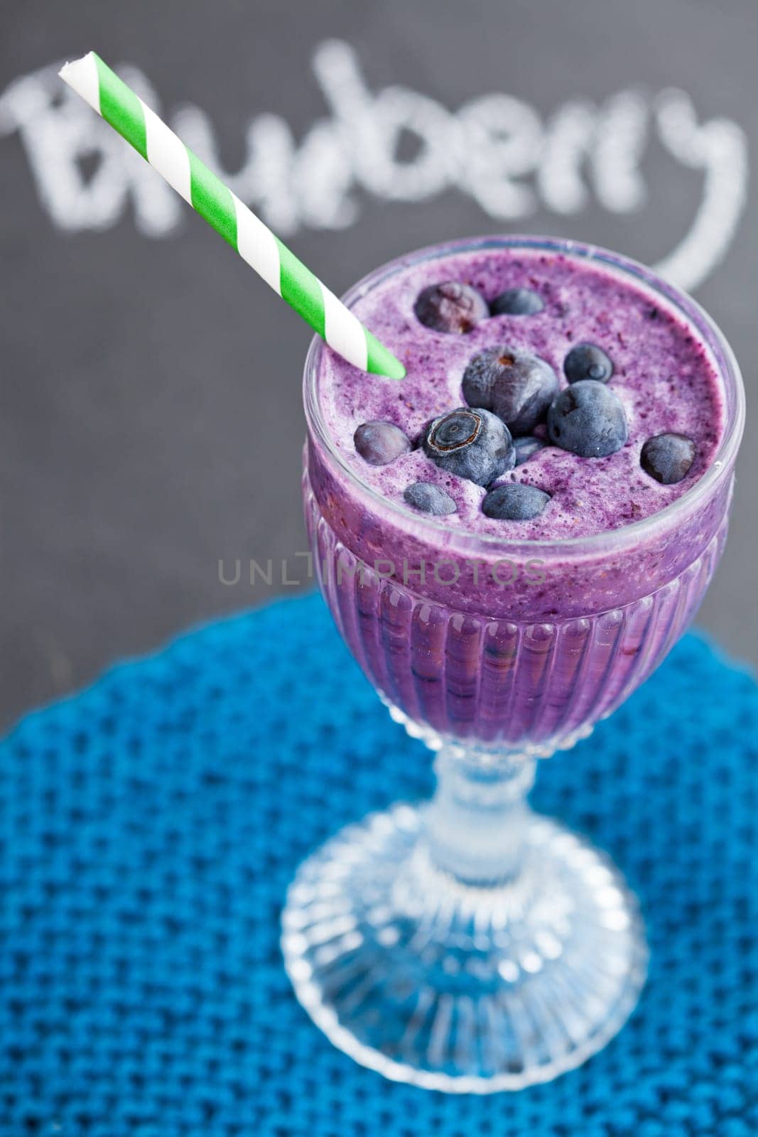 Closeup of blueberry milk smoothie in glass with striped straw on blue mat with word blueberry written on dark background