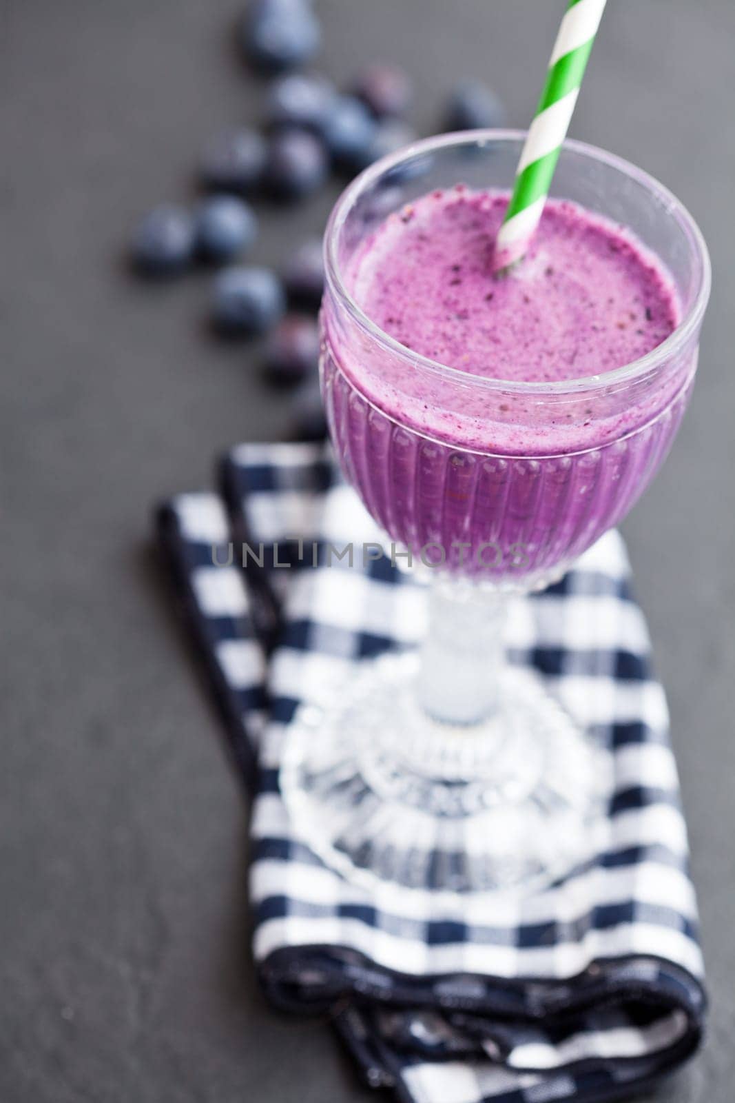 Closeup of blueberry milk smoothie in glass with striped straw on checkered napkin and fresh blueberries in background