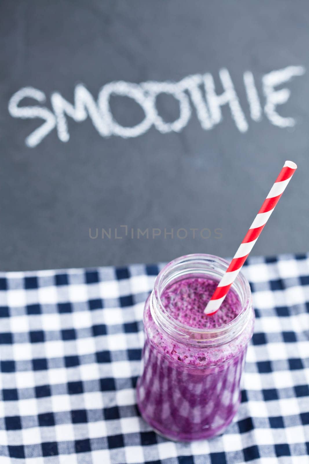 Closeup of blueberry smoothie in glass with striped straw resting on checkered napkin with word smoothie written in chalk on dark background