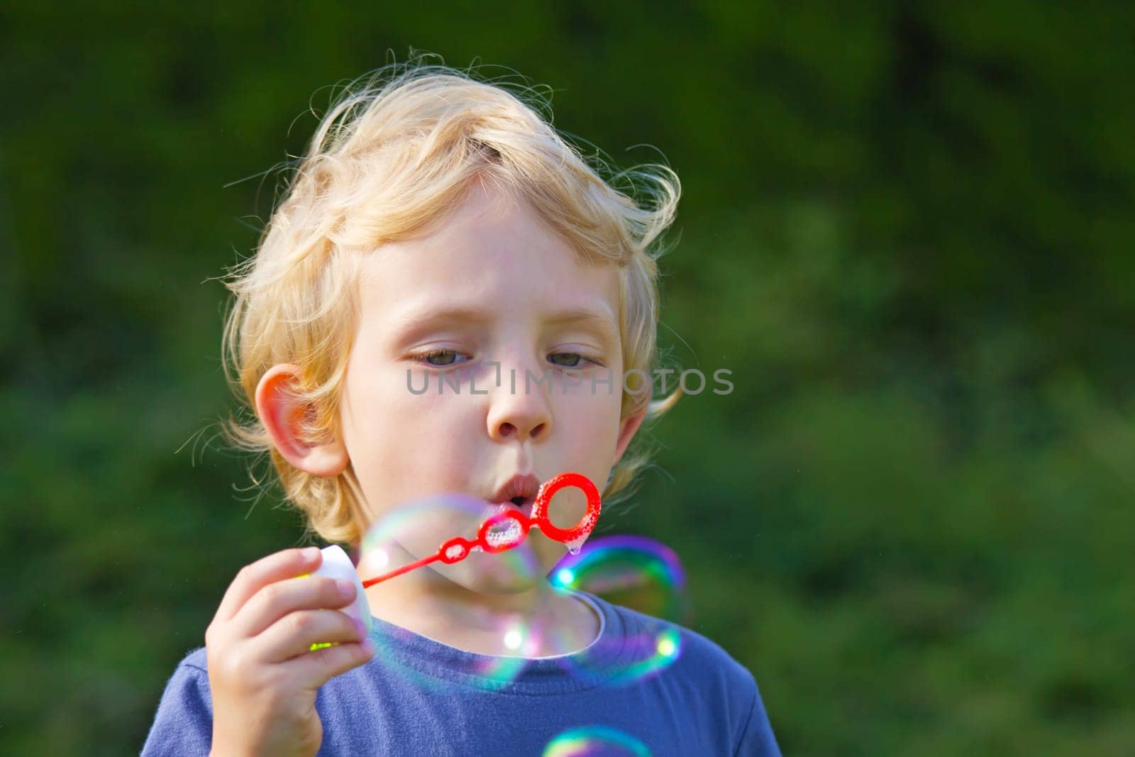 Blond boy making soap bubbles outside (green background)