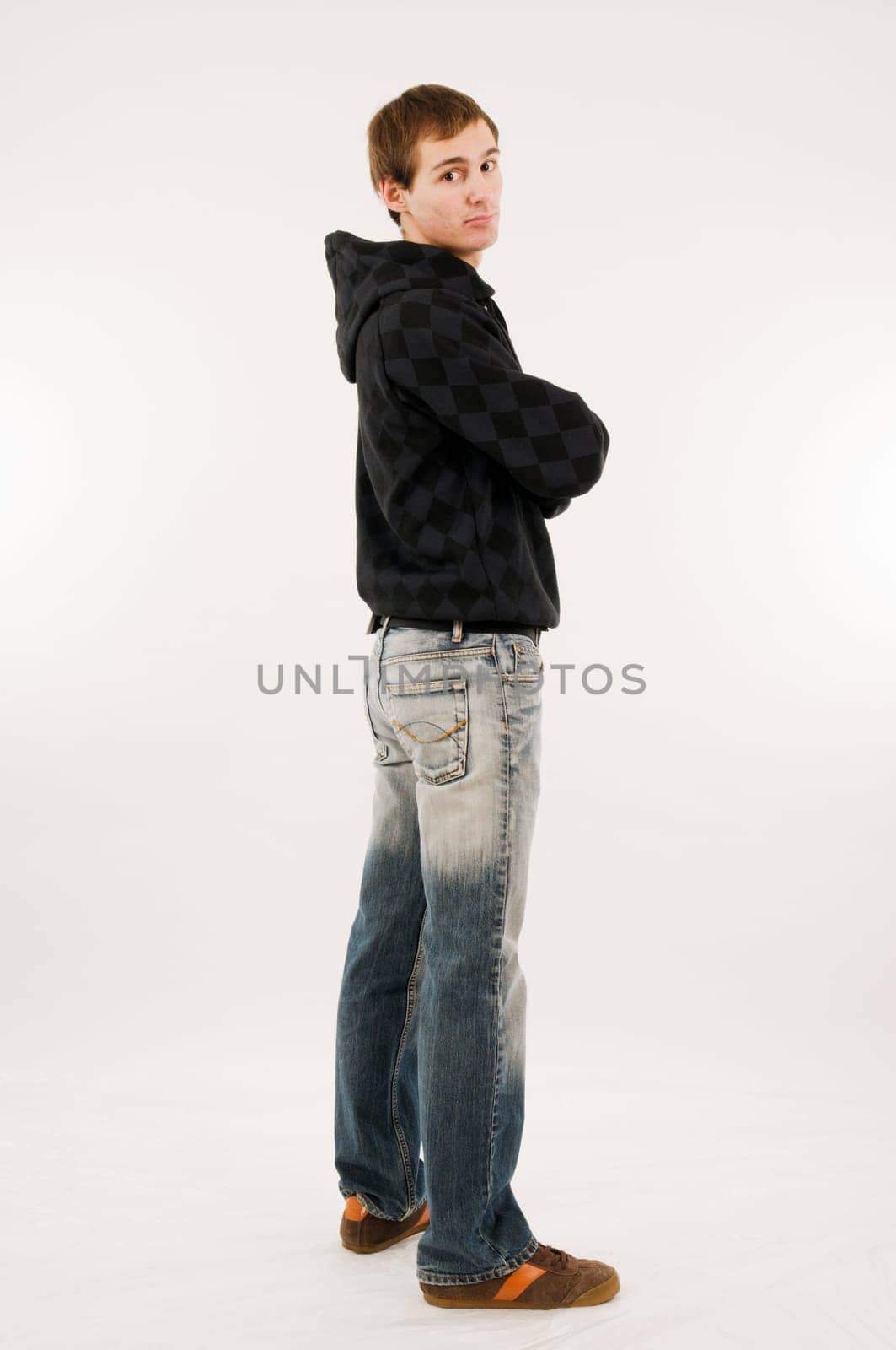 Side full body shot of a young man in casual casual clothes with crossed arms over his shoulder looking seriously into the camera against a white background