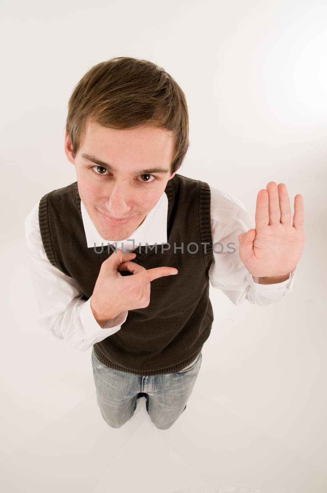 wide-angle top view of a young man in front of a white background in a white shirt and brown vestover  the hand raised in oath,looking into the camera and pointing with the right hand grinning to the left