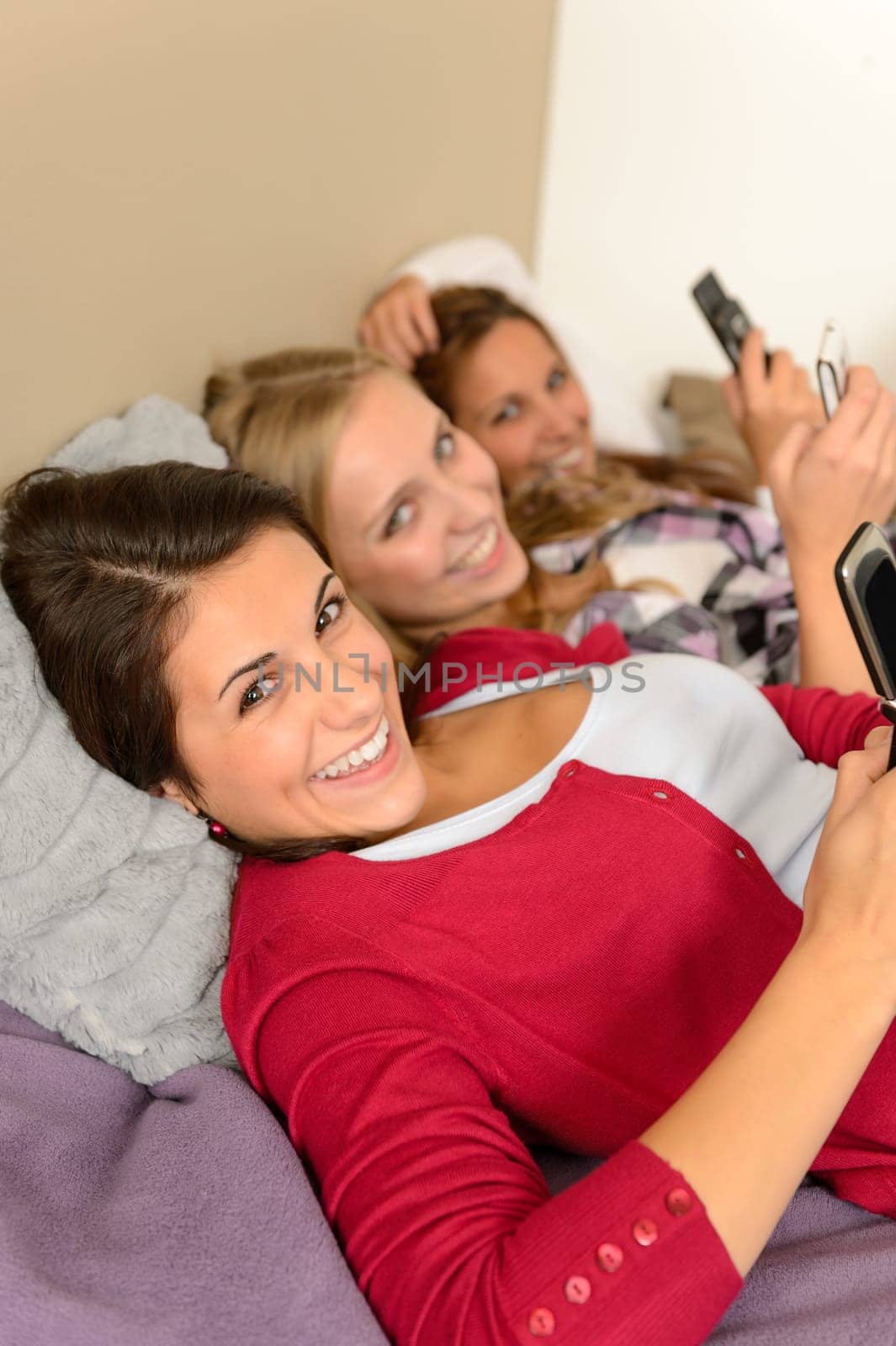 Three smiling young girl lying on bed using smart phones