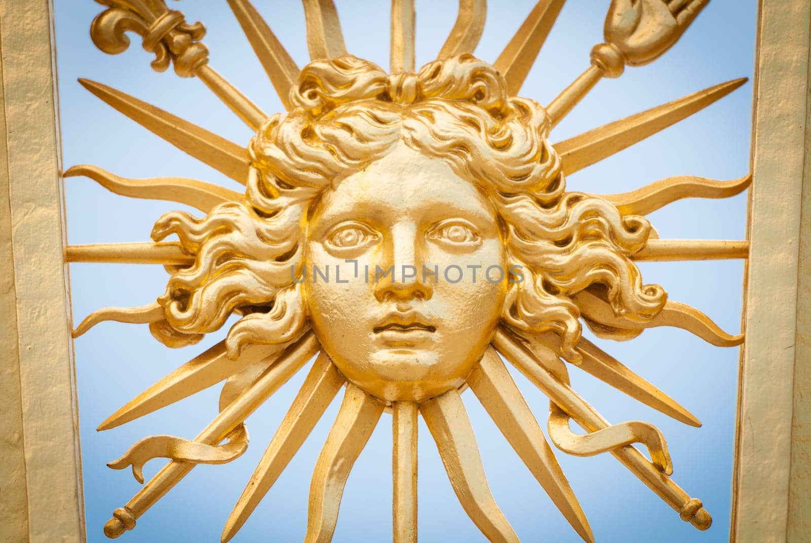 Detail view of golden ornate gate of Chateau de Versailles depicting human face. Blue sky in background. Paris, France, Europe.