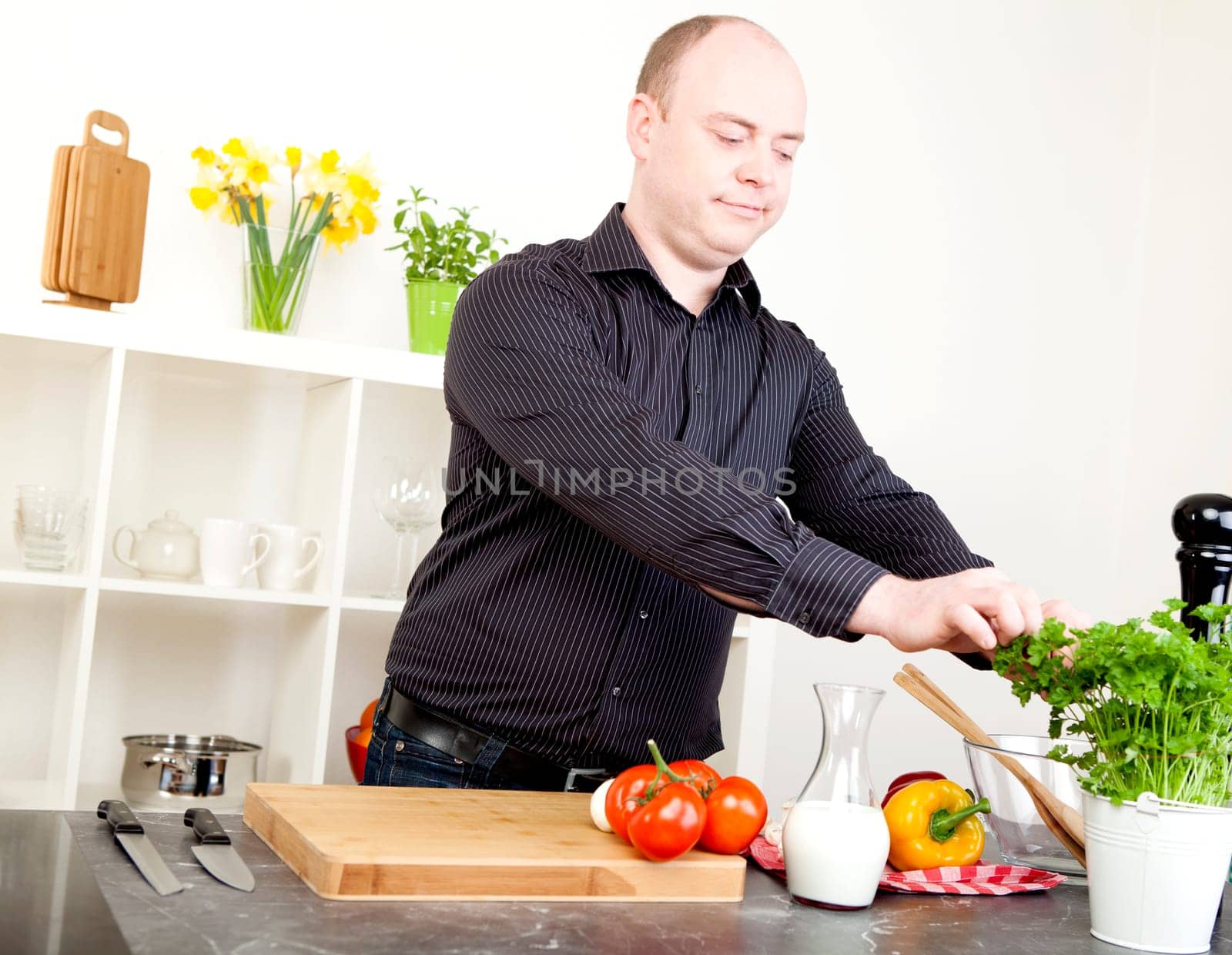 Man picking fresh herbs from a potted plant while standing in a clean white kitchen preparing a meal with fresh vegetables