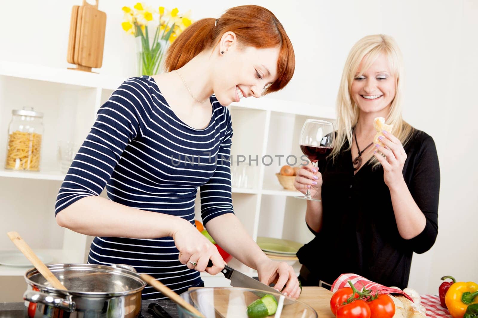 Two attractive women friends preparing a meal in the kitchen together with one chopping vegetables watched by the second who is enjoying a glass of red wine