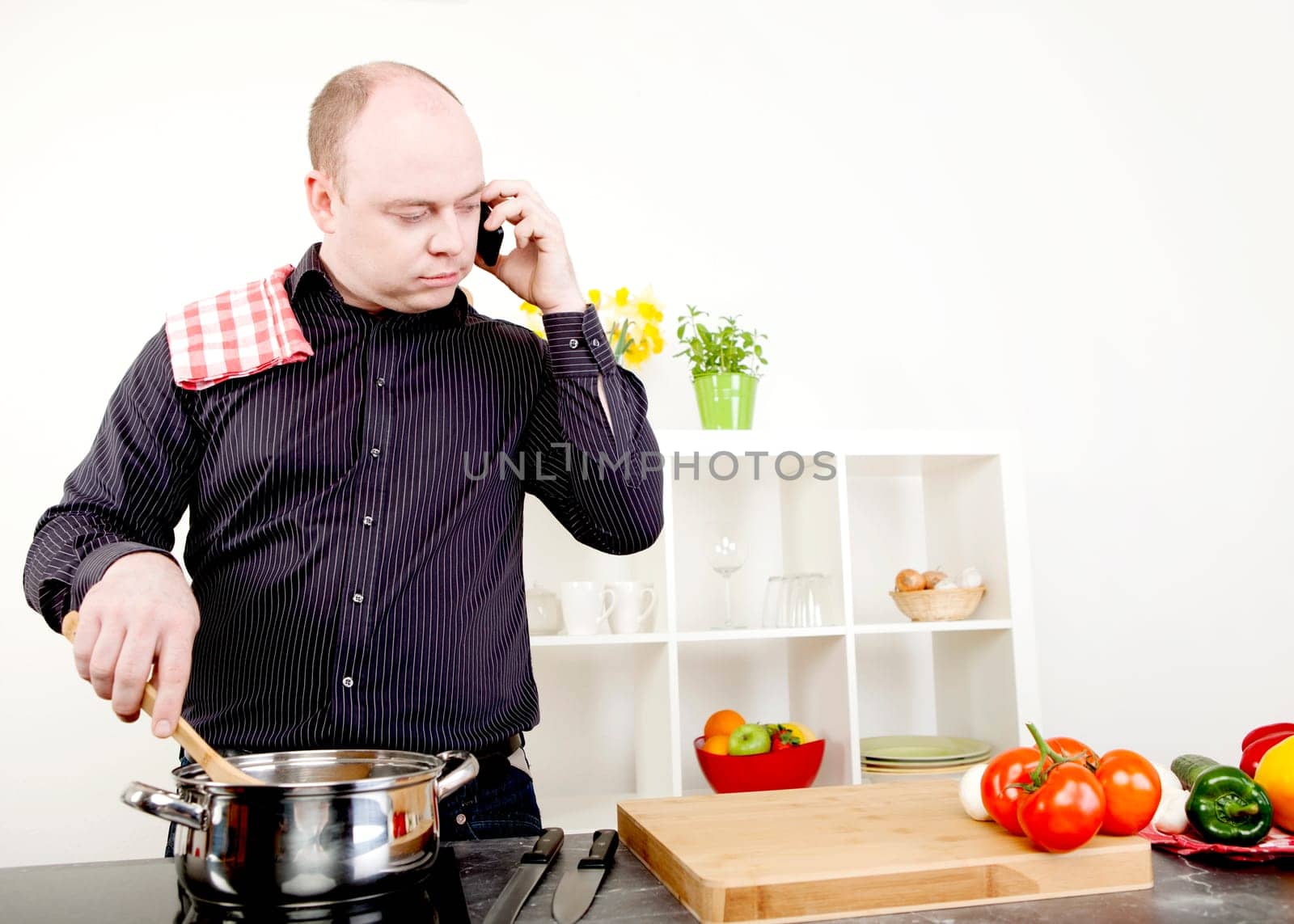 Attractive middle-aged balding man taking a call on his mobile while standing cooking at the stove stirring the contents of a stainless steel saucepan