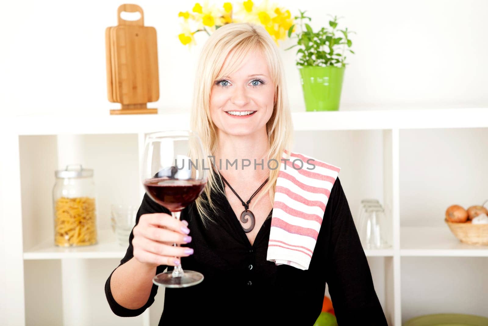 Elegant beautiful blonde woman standing in the kitchen with a dishcloth over her shoulder smiling and making a toast with a glass of red wine