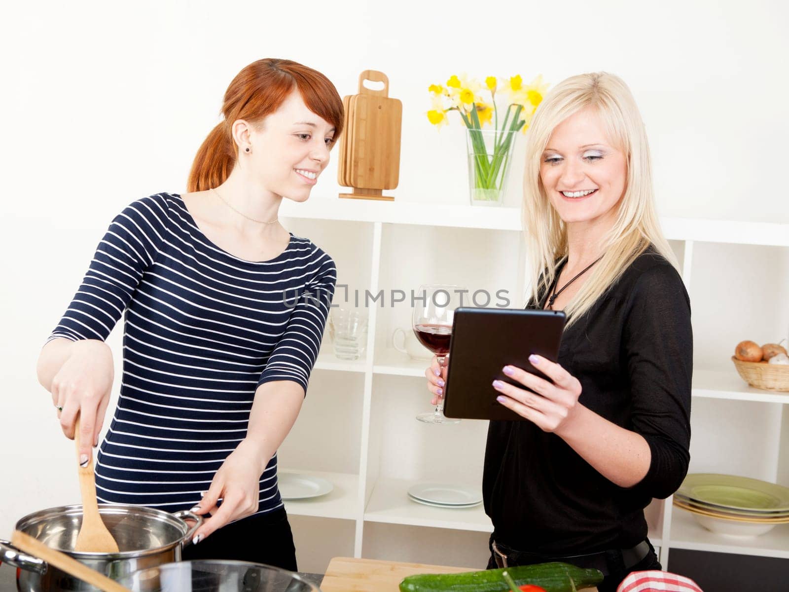 Two attractive young female friends cooking in the kitchen reading the screen on a tablet while one stands stirring a saucepan on the stove