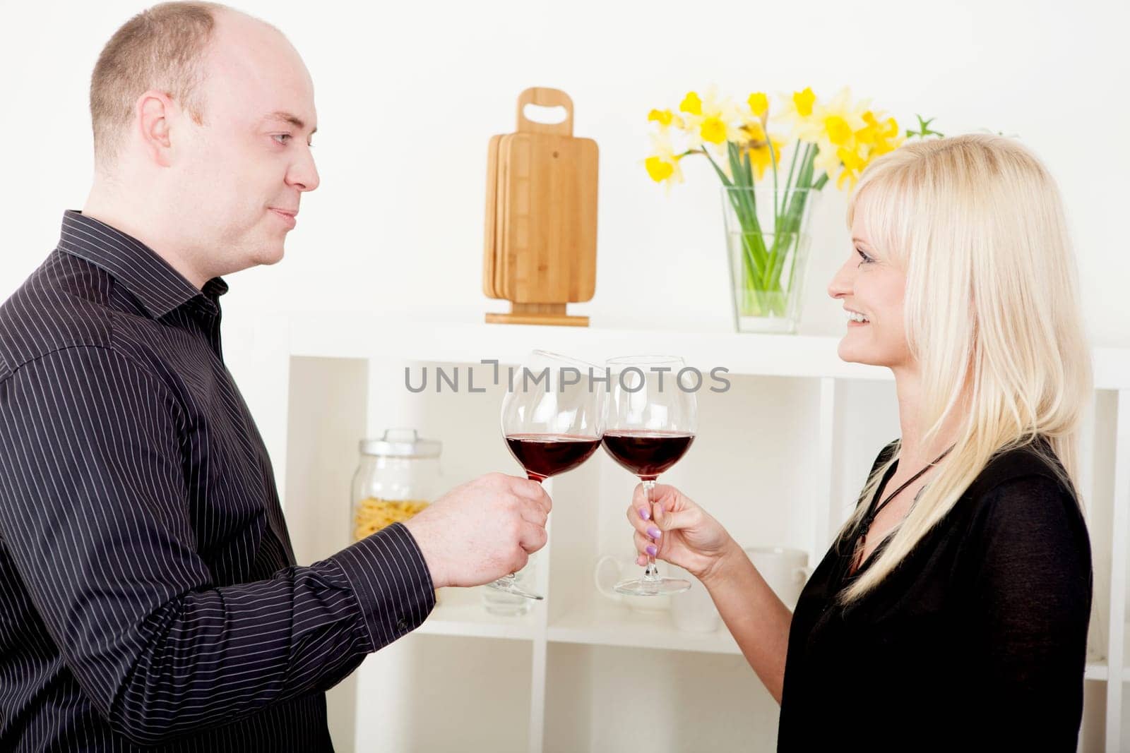 Romantic attractive man and woman standing sideways to the camera clinking their glasses of red wine in celebration toasting each other while looking into each others eyes with a smile