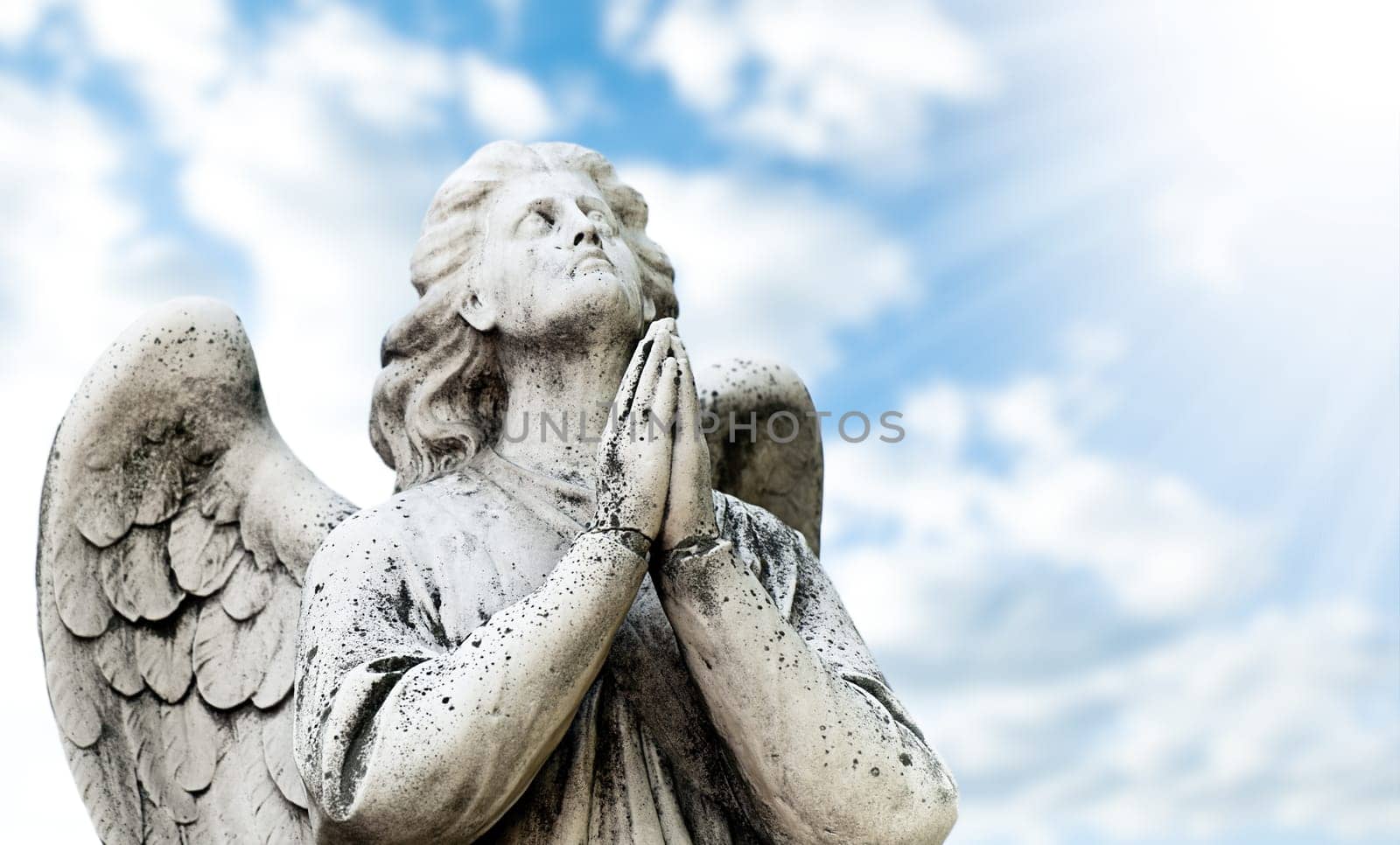 Beautiful statue of the angel praying, blue sky with clouds and sun with rays in background