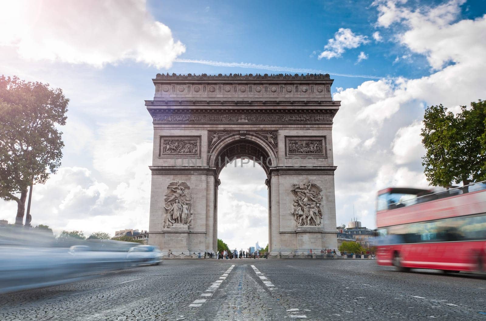 Triumphal arch. Blue sky and white clouds in background. Blurred cars and red tourist bus in foreground. Paris, France, Europe.