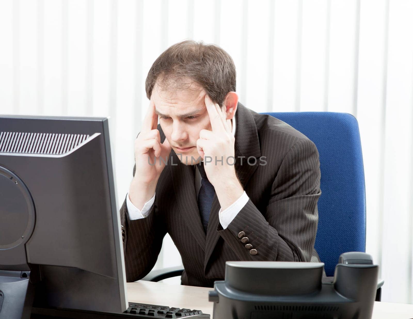 Worried businessman with a headache sitting at his desk rubbing his temples with his fingertips and staring at his desktop computer monitor in despair