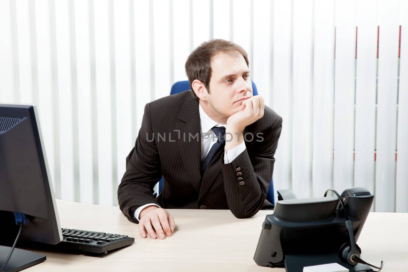 Thoughtful businessman in his office sitting at his desk resting his chin on his hand as he stares into the distance while contemplating difficult decisions