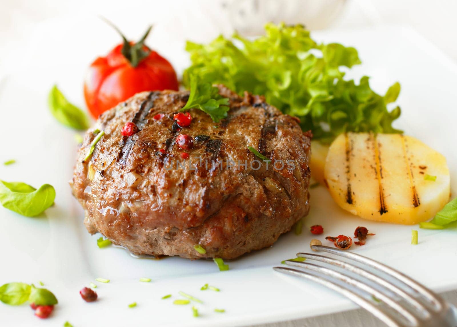 Delicious grilled beef meatball served on a white plate with tomato and lettuce with a fork in the foreground