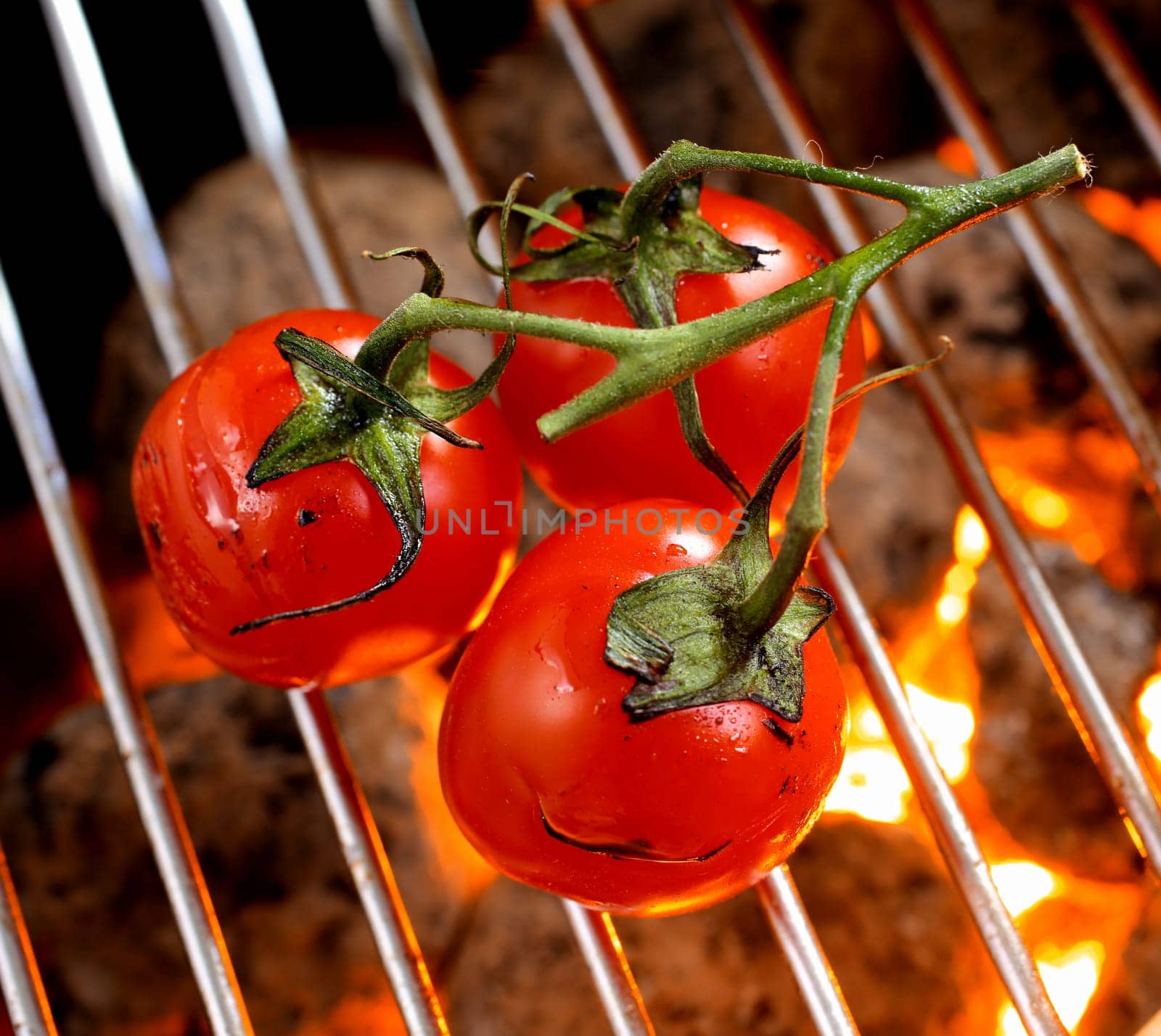 Bunch of ripe red cherry tomatoes on the vine roasting over a barbecue fire, high angle view of the grid and glowing charcoal