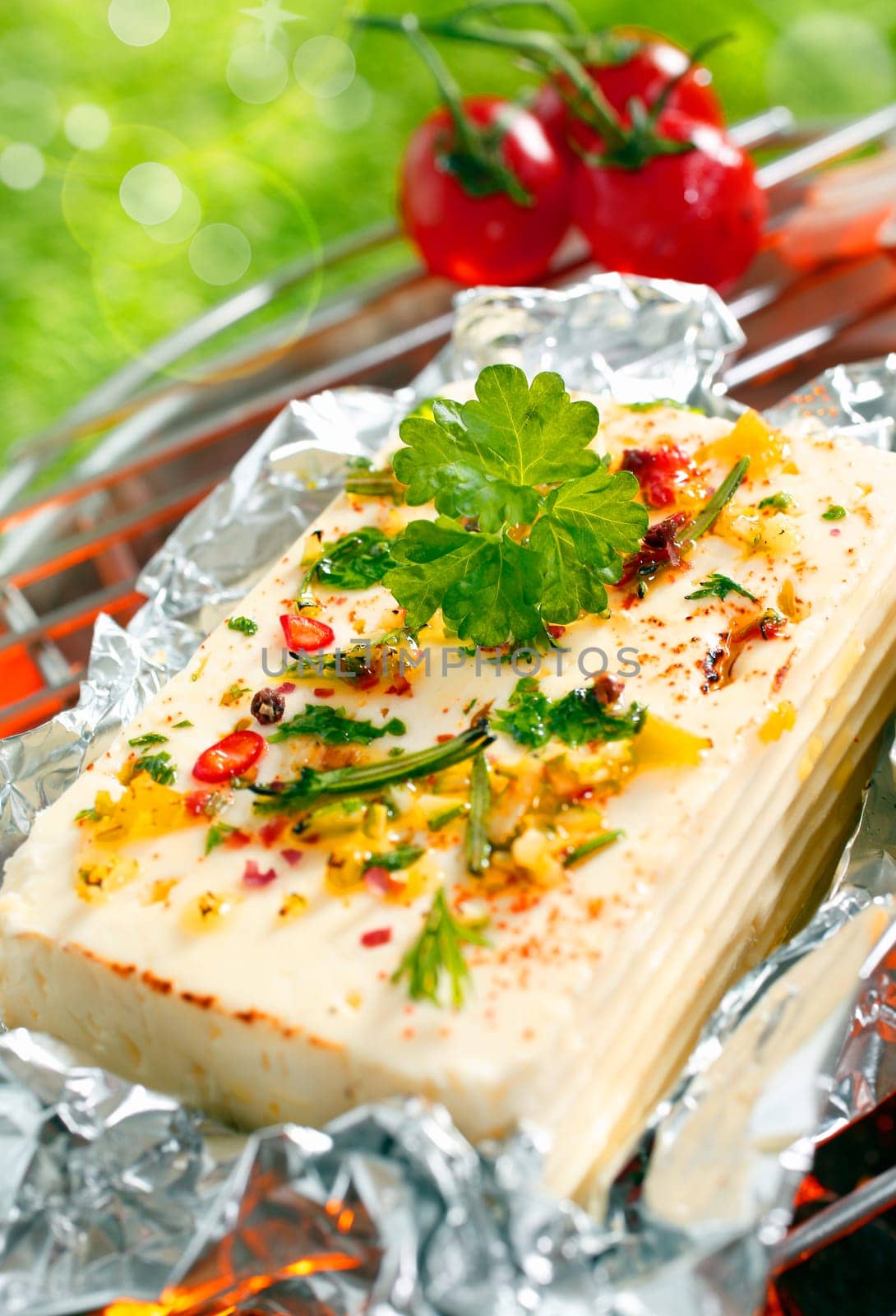 Halloumi cheese topped with fresh herbs and spices grilling in tin foil over the glowing coals of a barbecue outdoors in the sunshine