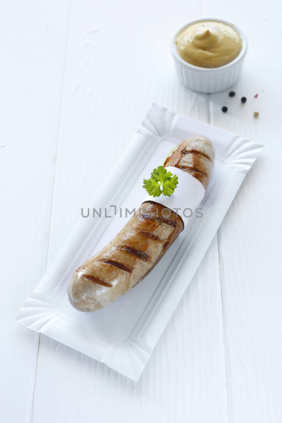 One cooked sausage with a sprig of flat parsley on a white plate with a pot of mustard.