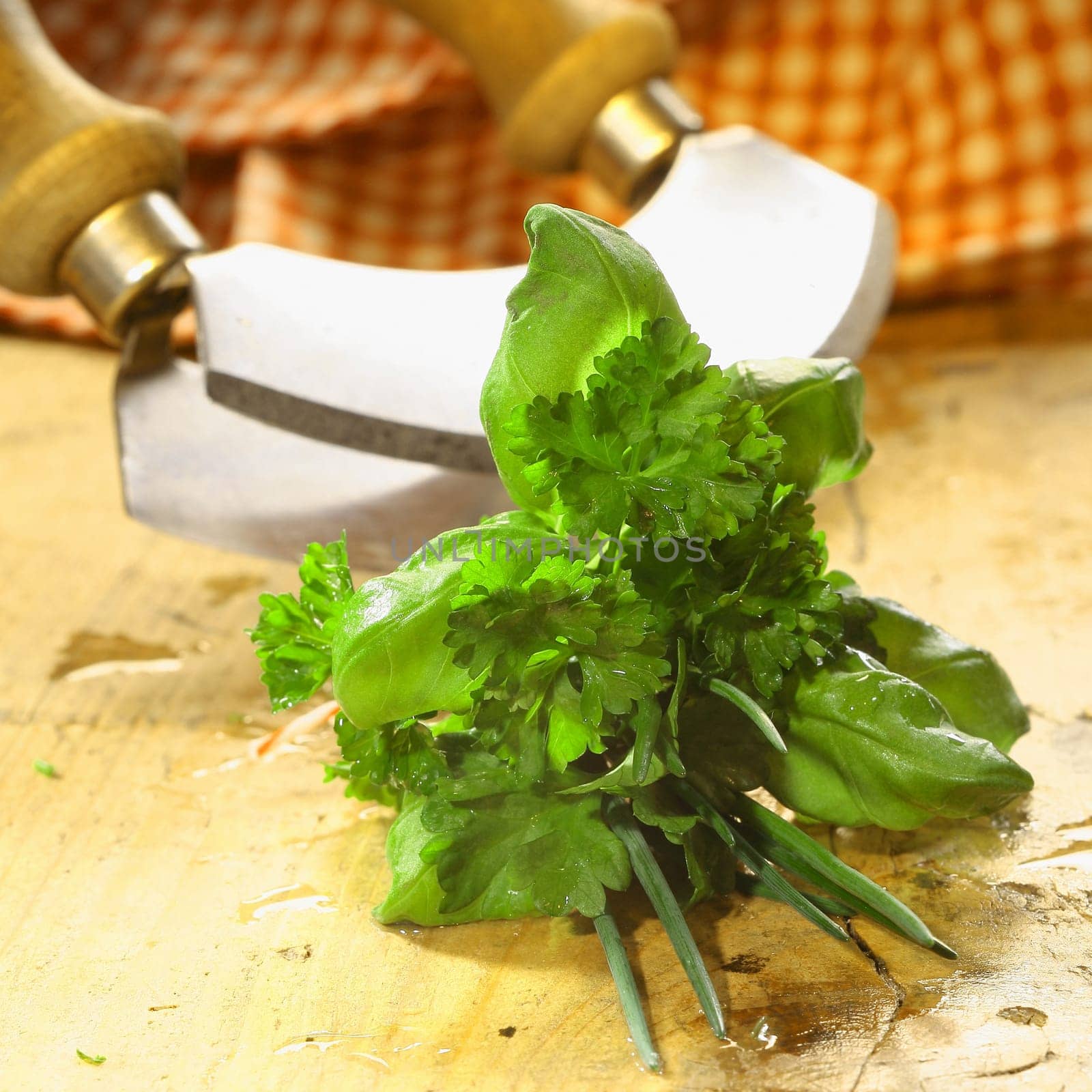 Bouquet garni of fresh herbs including basil, crinkly leaf parsley and chives lying on a wooden table in front of a chopping blade