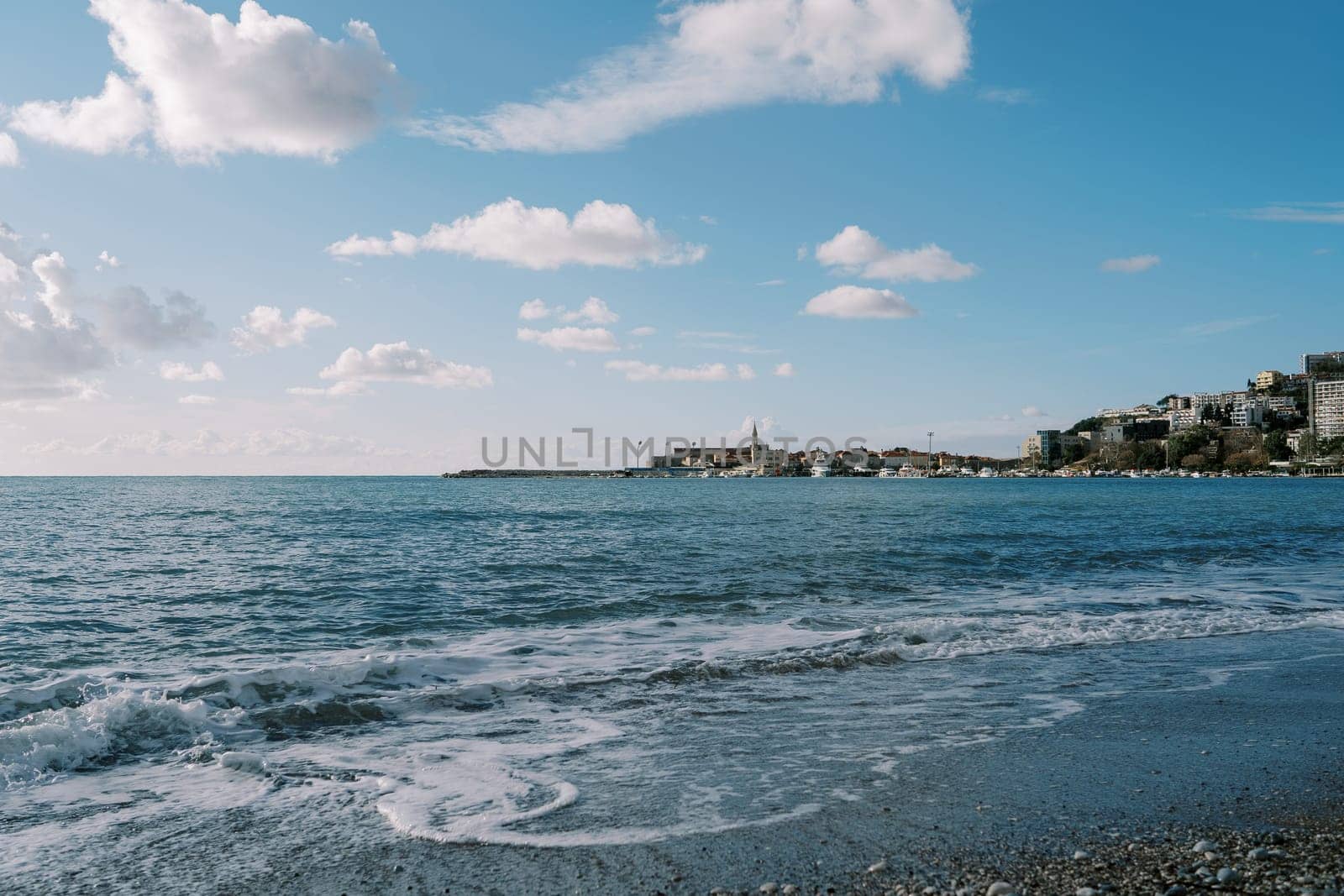 Sea surf on the beach against the backdrop of the resort town and cloudy sky by Nadtochiy