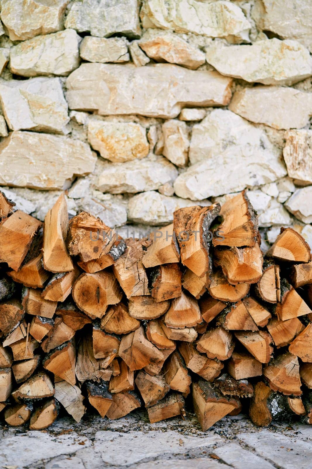 Stack of firewood is stacked against the stone wall of a house on a paved path. High quality photo