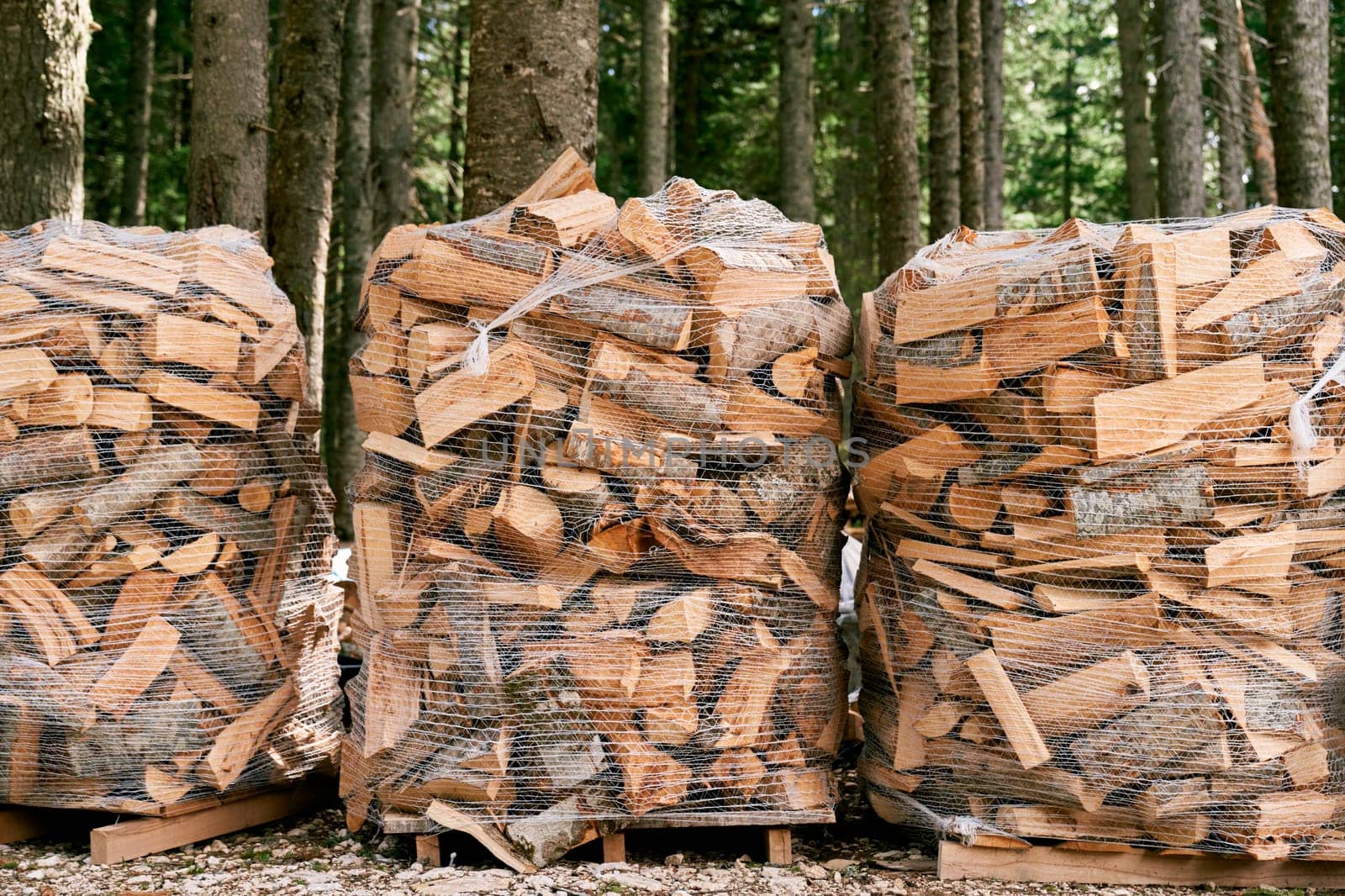 Stacks of firewood lie in transparent mesh bags on pallets among the trees. High quality photo