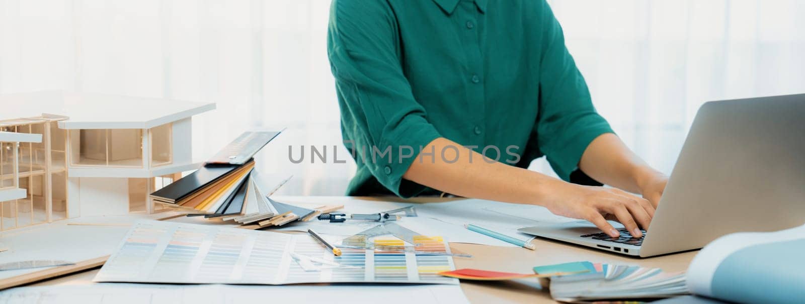 Cropped image of professional interior designer using laptop to create house plan on table with color palette and architectural model scatter around. Creative working and design concept. Variegated.