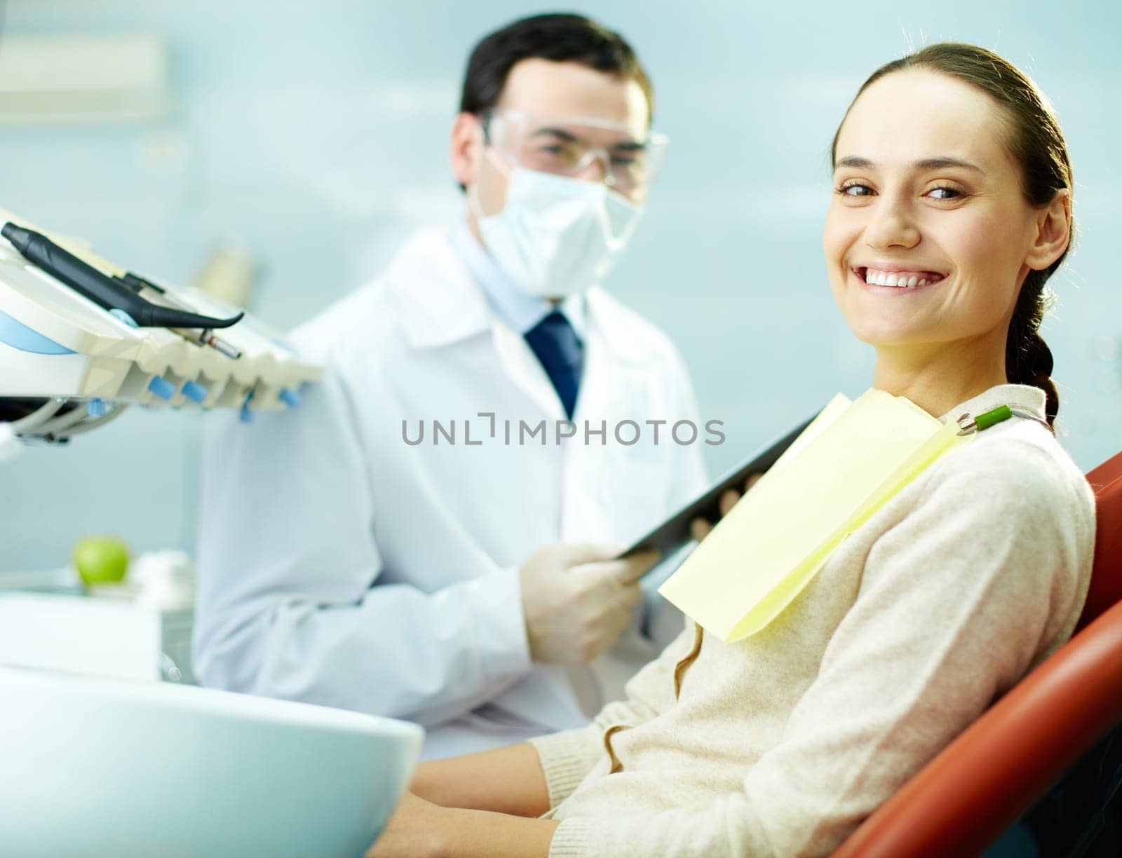 Smiling woman sitting in dentists chair with doctor on background