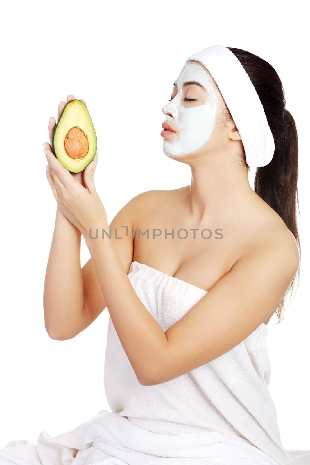 Young woman holding avocado heaving face clay mask on the face in a spa