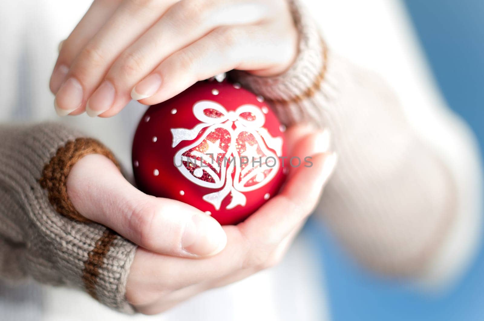 young woman against blue background wearing winter clothes holding red christmas ball toy in her hands