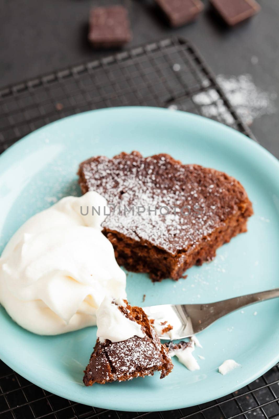 Closeup of slice of chocolate cake and whipped cream on plate resting on mesh metal tray with chocolate squares in background