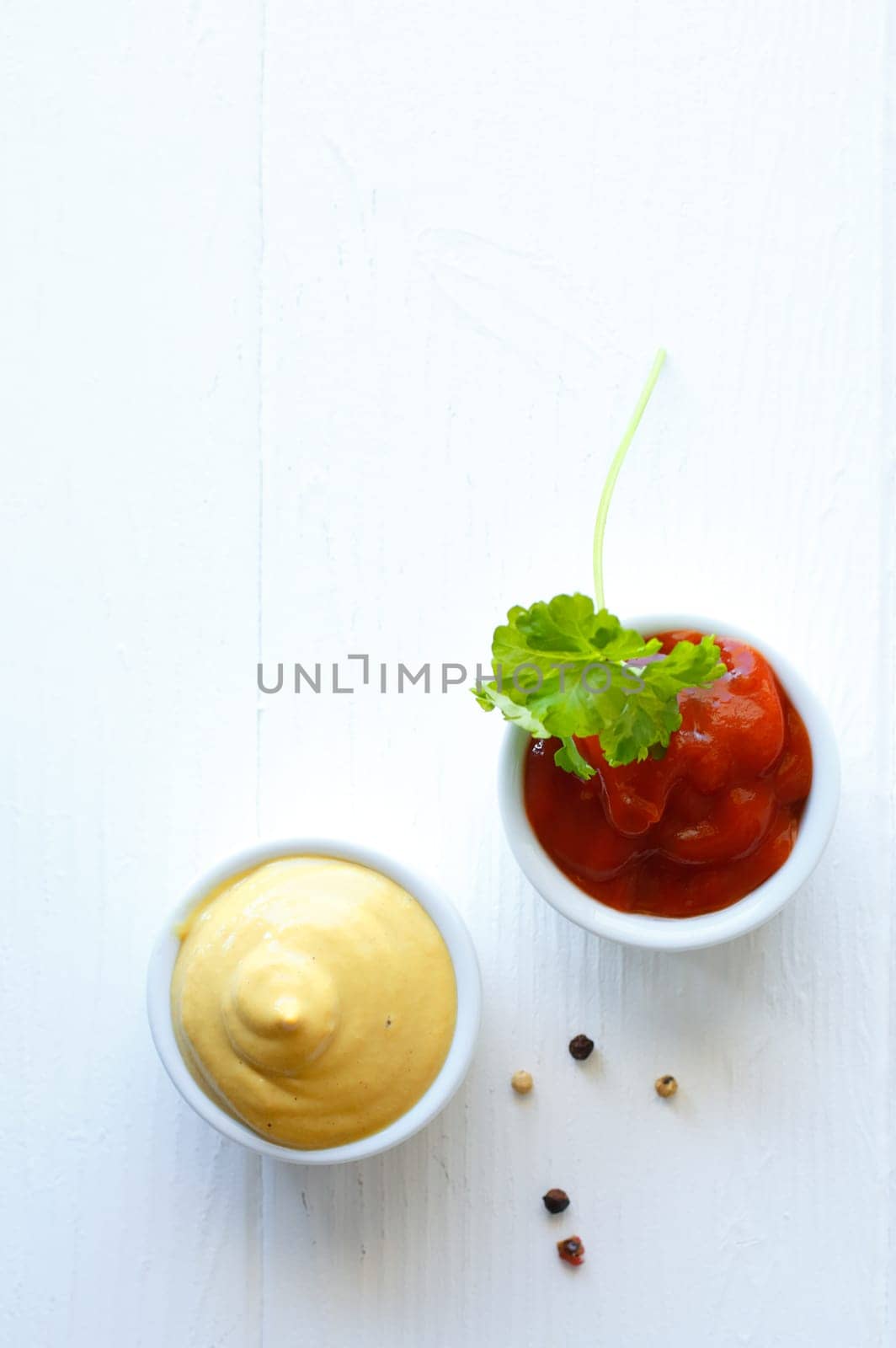 Overhead view of servings of mustard and tomato ketchup in small individual ceramic ramekins garnished with parsley on a white background with copyspace