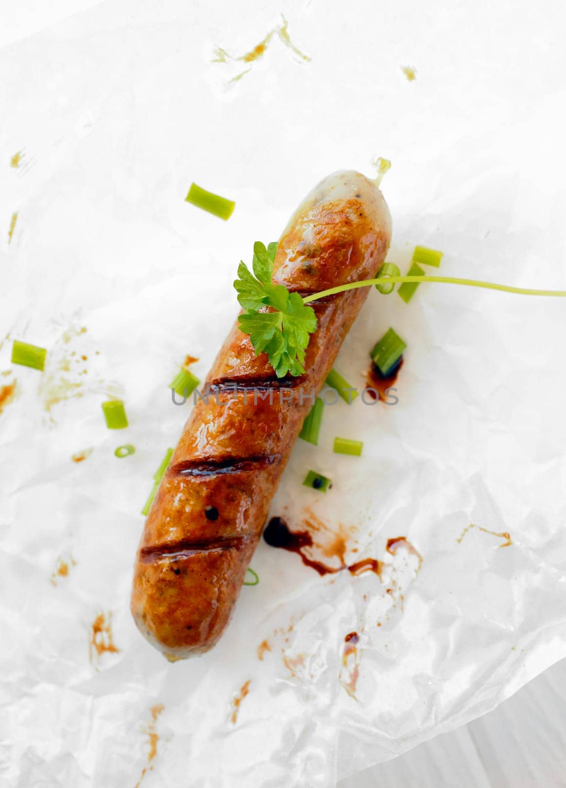 Bird's eye view of one cooked sausage on a plate with a sprig of parsley and chopped spring onions.