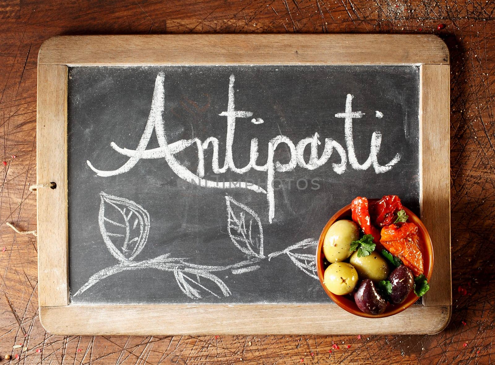 Chalkboard Antipasti sign with handwritten text and decorative foliage with a small bowl of black and green olives and hot chilli peppers in the corner, overhead view on a wooden background